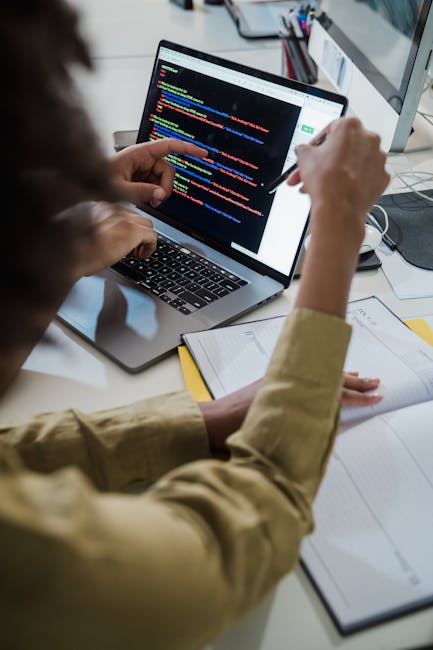 A person using a laptop on a desk, viewing a screen displaying a code editor with colorful programming lines. Their right hand holds a pen, indicating they may be making notes or modifications. The de