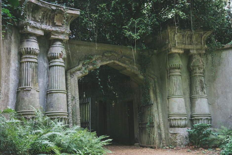An ancient stone garden entrance framed by classical-style columns and a carved archway over an iron gate, partially obscured by lush green foliage and creeping vines. The weathered stone features mos