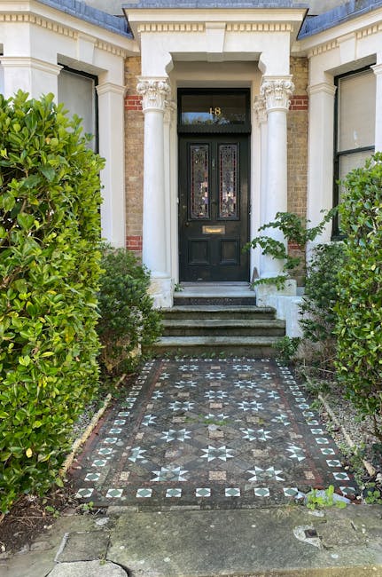 An ancient stone garden entrance framed by classical-style columns and a carved archway over an iron gate, partially obscured by lush green foliage and creeping vines. The weathered stone features mos